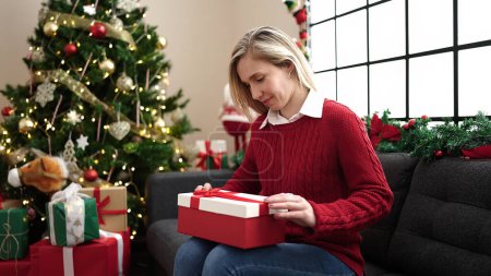 Foto de Joven mujer rubia desembalaje regalo sentado en el sofá por el árbol de Navidad en casa - Imagen libre de derechos