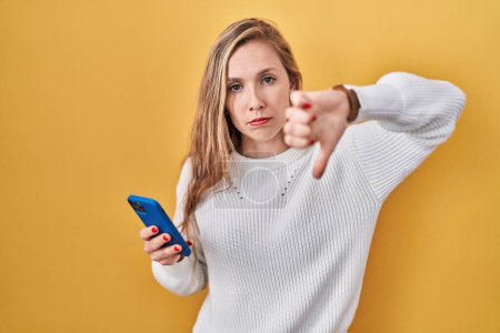 Photo for Young blonde woman using smartphone typing message looking unhappy and angry showing rejection and negative with thumbs down gesture. bad expression. - Royalty Free Image