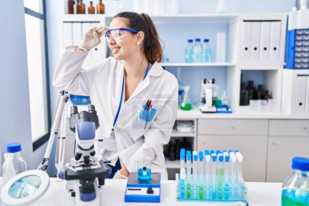 Photo for Young hispanic woman working at scientist laboratory very happy and smiling looking far away with hand over head. searching concept. - Royalty Free Image