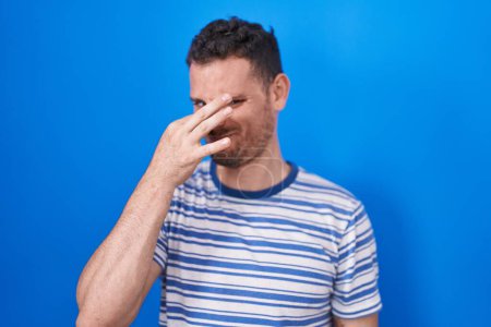 Photo for Young hispanic man standing over blue background smelling something stinky and disgusting, intolerable smell, holding breath with fingers on nose. bad smell - Royalty Free Image