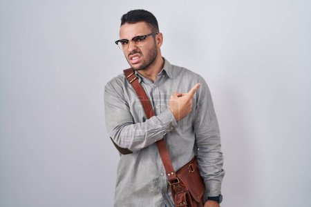 Téléchargez les photos : Jeune homme hispanique portant une valise pointant de côté inquiet et nerveux avec index, préoccupé et expression surprise - en image libre de droit
