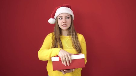 Téléchargez les photos : Jeune femme blonde portant un chapeau de Noël tenant un cadeau sur fond rouge isolé - en image libre de droit