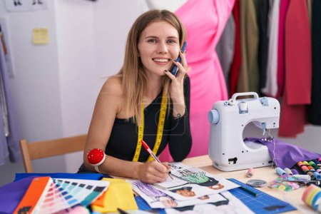 Téléchargez les photos : Jeune femme blonde tailleur parler sur smartphone dessin conception de vêtements au magasin de tailleur - en image libre de droit