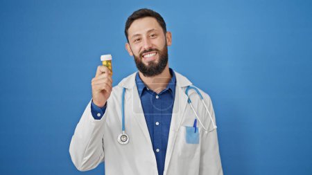 Téléchargez les photos : Jeune homme hispanique médecin tenant bouteille de pilules sur fond bleu isolé - en image libre de droit