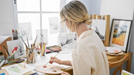 Téléchargez les photos : Jeune artiste blonde dessinant sur du papier assis sur une table au studio d'art - en image libre de droit