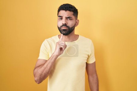Photo for Hispanic man with beard standing over yellow background thinking concentrated about doubt with finger on chin and looking up wondering - Royalty Free Image