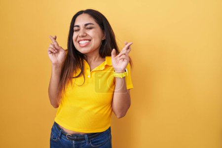 Téléchargez les photos : Jeune femme arabe debout sur fond jaune geste doigt croisé souriant avec espoir et les yeux fermés. chance et concept superstitieux. - en image libre de droit