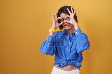 Photo for Young girl standing over yellow background doing ok gesture like binoculars sticking tongue out, eyes looking through fingers. crazy expression. - Royalty Free Image
