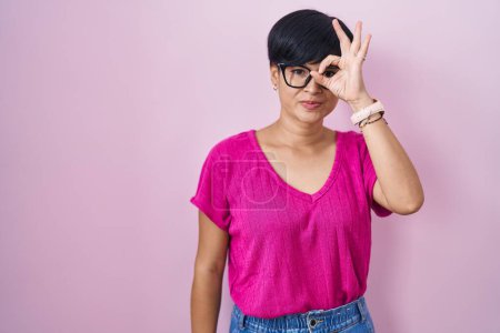 Foto de Joven mujer asiática con pelo corto de pie sobre fondo rosa haciendo buen gesto con la mano sonriente, ojos mirando a través de los dedos con la cara feliz. - Imagen libre de derechos