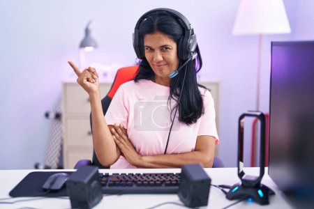 Téléchargez les photos : Femme hispanique mature jouant à des jeux vidéo à la maison avec un grand sourire sur le visage, pointant avec la main et le doigt sur le côté en regardant la caméra. - en image libre de droit
