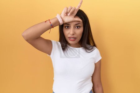 Téléchargez les photos : Jeune femme arabe portant un t-shirt blanc décontracté sur fond jaune se moquant des gens avec les doigts sur le front faisant geste de perdant se moquant et insultant. - en image libre de droit