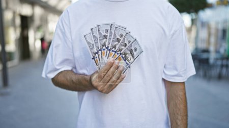 Photo for Hispanic man's hands casually holding dollar bills on a city street, a symbolic act of urban finance and investment in the vibrant outdoor sunlight. - Royalty Free Image