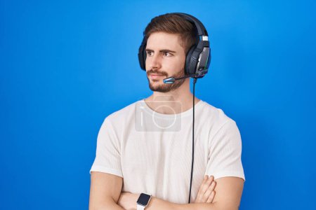 Téléchargez les photos : Homme hispanique avec barbe écoutant de la musique portant des écouteurs regardant sur le côté avec les bras croisés convaincu et confiant - en image libre de droit