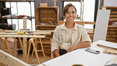 Fröhliche junge hispanische Tischlerin sitzt am Tisch und lächelt selbstbewusst in ihrer Tischlerwerkstatt