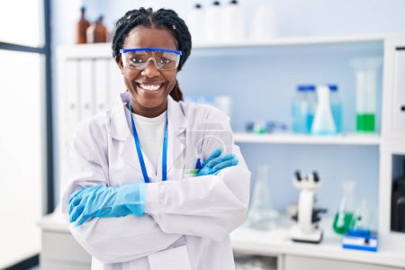 Foto de Mujer científica afroamericana sonriendo confiada de pie con los brazos cruzados gesto en el laboratorio - Imagen libre de derechos