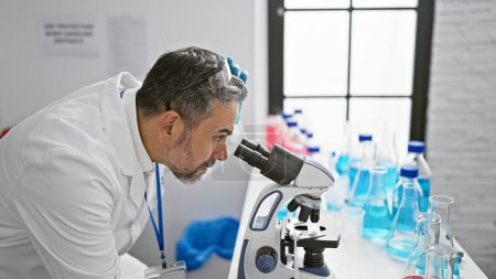 Photo for Attractive grey-haired young hispanic man, a dedicated scientist, working intently on a medical experiment in the lab, safety gloves on, peering through a microscope, deep in concentration. - Royalty Free Image