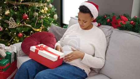 Photo for Young pregnant woman celebrating christmas holding gift at home - Royalty Free Image