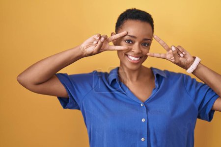 Téléchargez les photos : Femme afro-américaine debout sur fond jaune faisant symbole de paix avec les doigts sur le visage, souriant joyeux montrant la victoire - en image libre de droit
