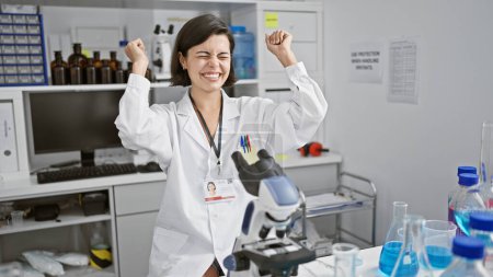 Téléchargez les photos : Célébrer une percée, jeune et belle scientifique hispanique, travaillant joyeusement avec un microscope dans le laboratoire, fait une découverte capitale - en image libre de droit