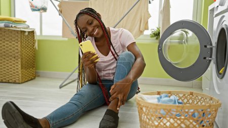 Téléchargez les photos : Belle femme afro-américaine assise sur le sol de la buanderie, souriant comme elle messages sur son smartphone au milieu des tâches domestiques - en image libre de droit
