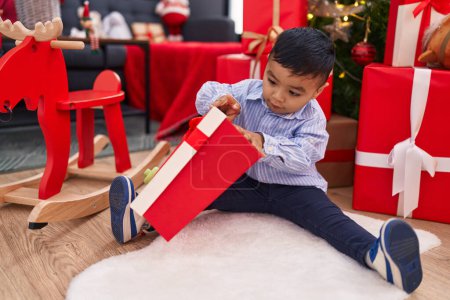 Foto de Adorable niño hispano desempacando regalo sentado junto al árbol de Navidad en casa - Imagen libre de derechos