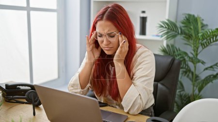 Photo for Stressed redhead young woman, an overworked business worker battling headache in office's hustle - Royalty Free Image