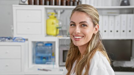 Téléchargez les photos : Une jeune scientifique blonde confiante exsudant son bonheur, souriante alors qu'elle se tient immergée dans une recherche médicale révolutionnaire, au milieu de l'agitation de son espace de travail de laboratoire animé. - en image libre de droit