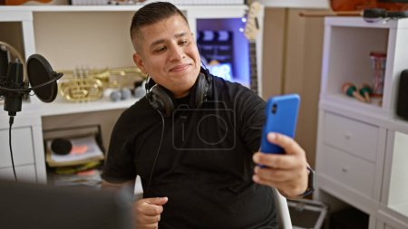 Foto de Guapo joven latino, un músico con auriculares, haciendo un selfie alegre con su teléfono inteligente dentro de un vibrante estudio de música. - Imagen libre de derechos
