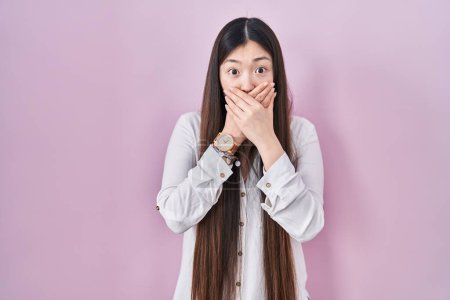 Foto de Mujer joven china de pie sobre el fondo rosa sorprendió cubriendo la boca con las manos por error. concepto secreto. - Imagen libre de derechos