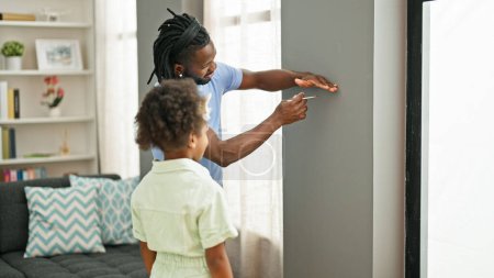 Foto de Padre e hija afroamericanos sonriendo confiados midiendo altura en la pared en casa - Imagen libre de derechos