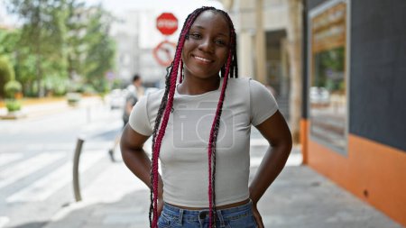 Téléchargez les photos : Belle femme afro-américaine riant, apportant de la joie dans la rue de la ville avec son sourire confiant et son style décontracté, rayonnant d'énergie positive sous le ciel ensoleillé - en image libre de droit
