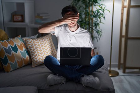 Photo for Young hispanic man using laptop at home at night smiling and laughing with hand on face covering eyes for surprise. blind concept. - Royalty Free Image