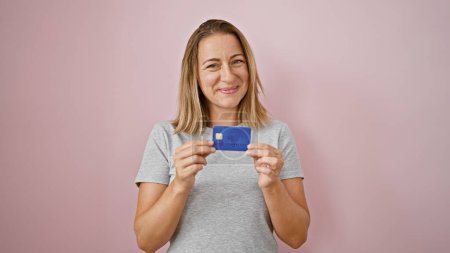 Photo for Cheerful young blonde woman confidently holds credit card, radiating joy and financial confidence over isolated pink background - Royalty Free Image