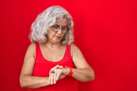 Téléchargez les photos : Femme d'âge moyen avec les cheveux gris debout sur fond rouge vérifier l'heure sur la montre-bracelet, détendu et confiant - en image libre de droit