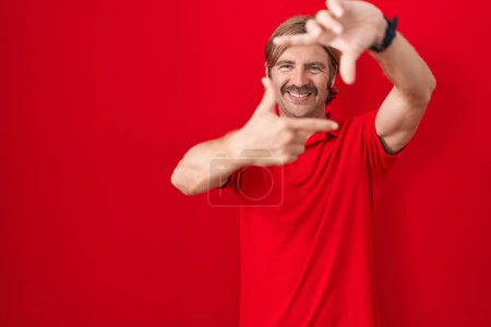 Téléchargez les photos : Homme caucasien avec moustache debout sur fond rouge souriant faisant cadre avec les mains et les doigts avec le visage heureux. concept de créativité et photographie. - en image libre de droit