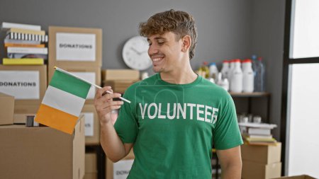 Téléchargez les photos : Jeune homme hispanique souriant faisant du bénévolat dans un centre caritatif, tenant fièrement le drapeau irlandais - en image libre de droit