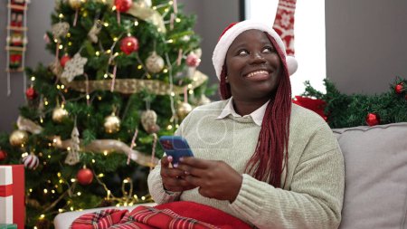 Téléchargez les photos : Femme africaine avec des cheveux tressés en utilisant un smartphone assis près de l'arbre de Noël à la maison - en image libre de droit