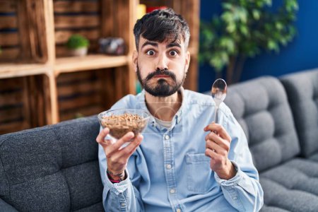 Téléchargez les photos : Jeune homme hispanique avec barbe mangeant des céréales à grains entiers en bonne santé soufflant des joues avec un drôle de visage. bouche gonflée d'air, attrapant l'air. - en image libre de droit