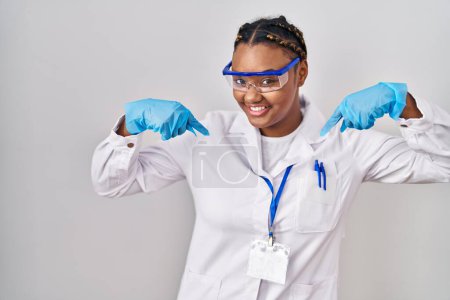 Foto de Mujer afroamericana con trenzas vestida con túnica científica que mira confiada con sonrisa en la cara, señalándose con los dedos orgullosos y felices. - Imagen libre de derechos