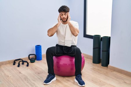 Téléchargez les photos : Homme hispanique avec barbe assis sur des boules de pilate à la salle de yoga avec la main sur la tête pour la douleur dans la tête parce que le stress. migraine souffrant. - en image libre de droit