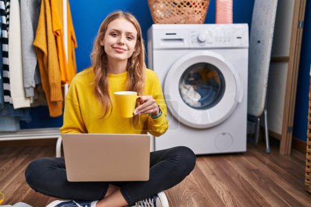 Téléchargez les photos : Jeune femme blonde utilisant un ordinateur portable buvant du café en attendant la machine à laver à la buanderie - en image libre de droit