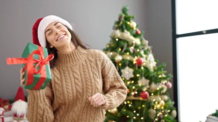 Foto de Joven mujer hispana hermosa sosteniendo regalo de Navidad en casa - Imagen libre de derechos