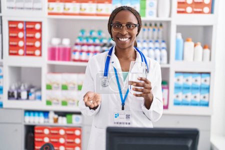 Foto de Farmacéutica afroamericana sosteniendo píldora y vaso de agua en farmacia - Imagen libre de derechos
