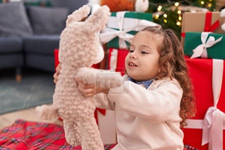 Téléchargez les photos : Adorable bambin blonde tenant ours en peluche assis sur le sol près de l'arbre de Noël à la maison - en image libre de droit