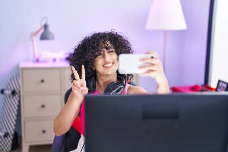 Téléchargez les photos : Jeune femme moyen-orientale streamer souriant confiant faire selfie par smartphone à la salle de jeu - en image libre de droit