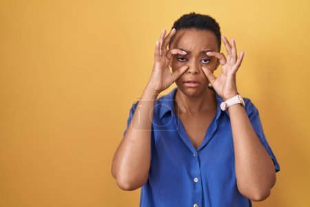 Photo for African american woman standing over yellow background trying to open eyes with fingers, sleepy and tired for morning fatigue - Royalty Free Image