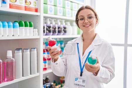 Téléchargez les photos : Jeune femme blonde pharmacienne souriante confiante tenant deux bouteilles à la pharmacie - en image libre de droit