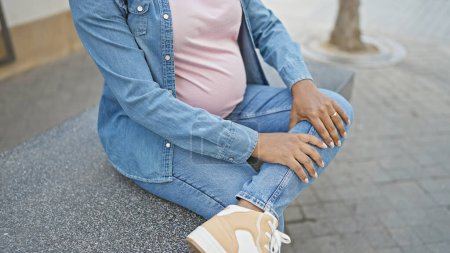 Téléchargez les photos : Jeune femme enceinte hispanique massant sa jambe fatiguée, assise sur un banc urbain à l'extérieur au soleil, irradiant une ambiance insouciante malgré les douleurs de la grossesse - en image libre de droit