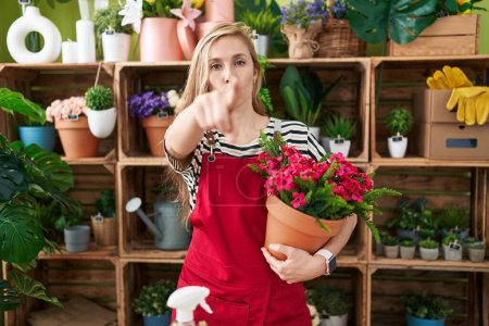 Foto de Joven mujer caucásica que trabaja en floristería sosteniendo la planta señalando con el dedo a la cámara y a usted, gesto de confianza mirando en serio - Imagen libre de derechos