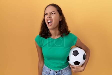 Foto de Mujer hispana joven sosteniendo la pelota enojada y loca gritando frustrada y furiosa, gritando con ira. rabia y concepto agresivo. - Imagen libre de derechos